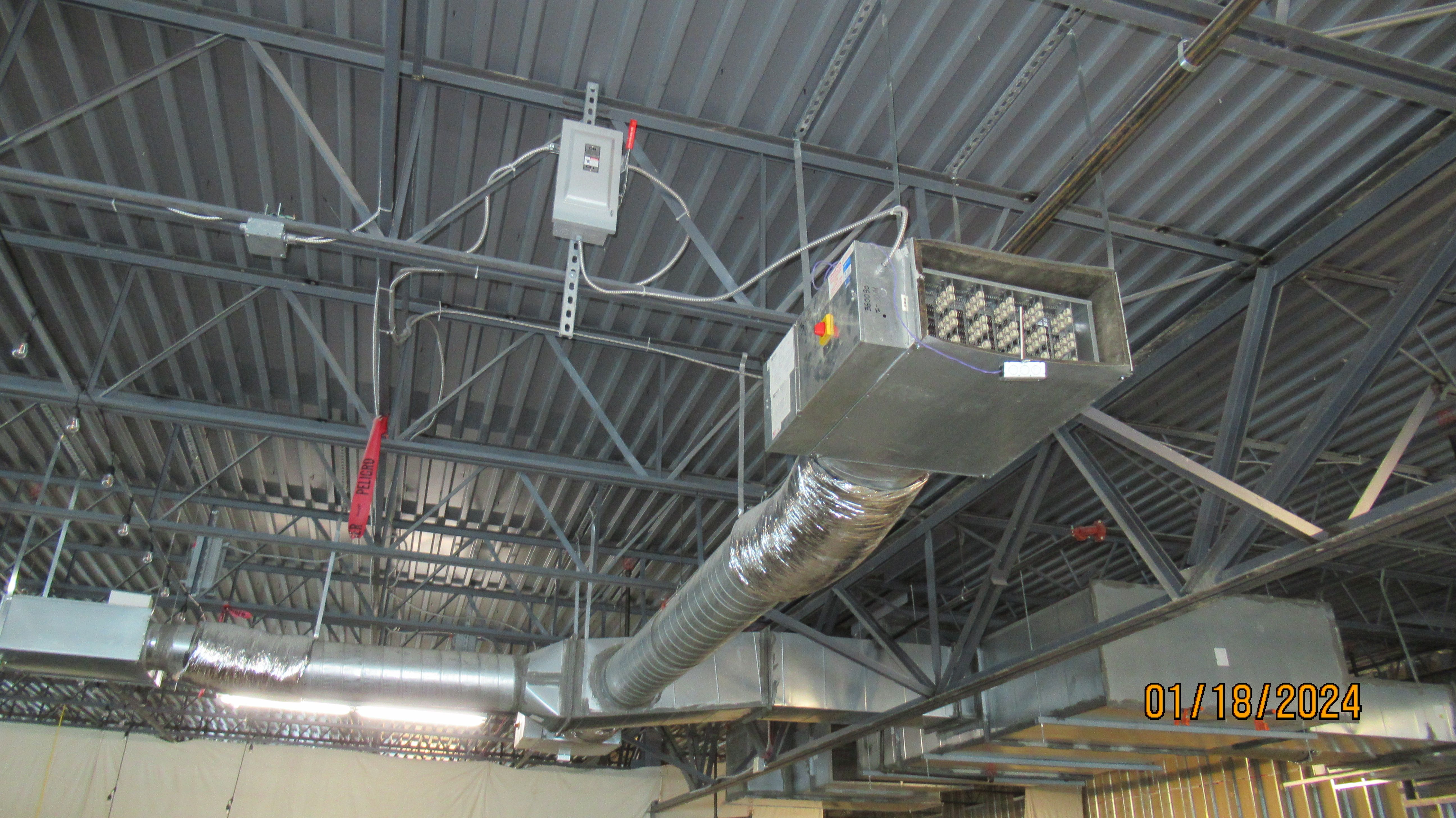 A suspended fan-coil air handler with an open coil section, connected to insulated flexible duct and galvanized sheet metal ductwork beneath an open steel roof deck, with an electrical disconnect switch mounted on a strut nearby.