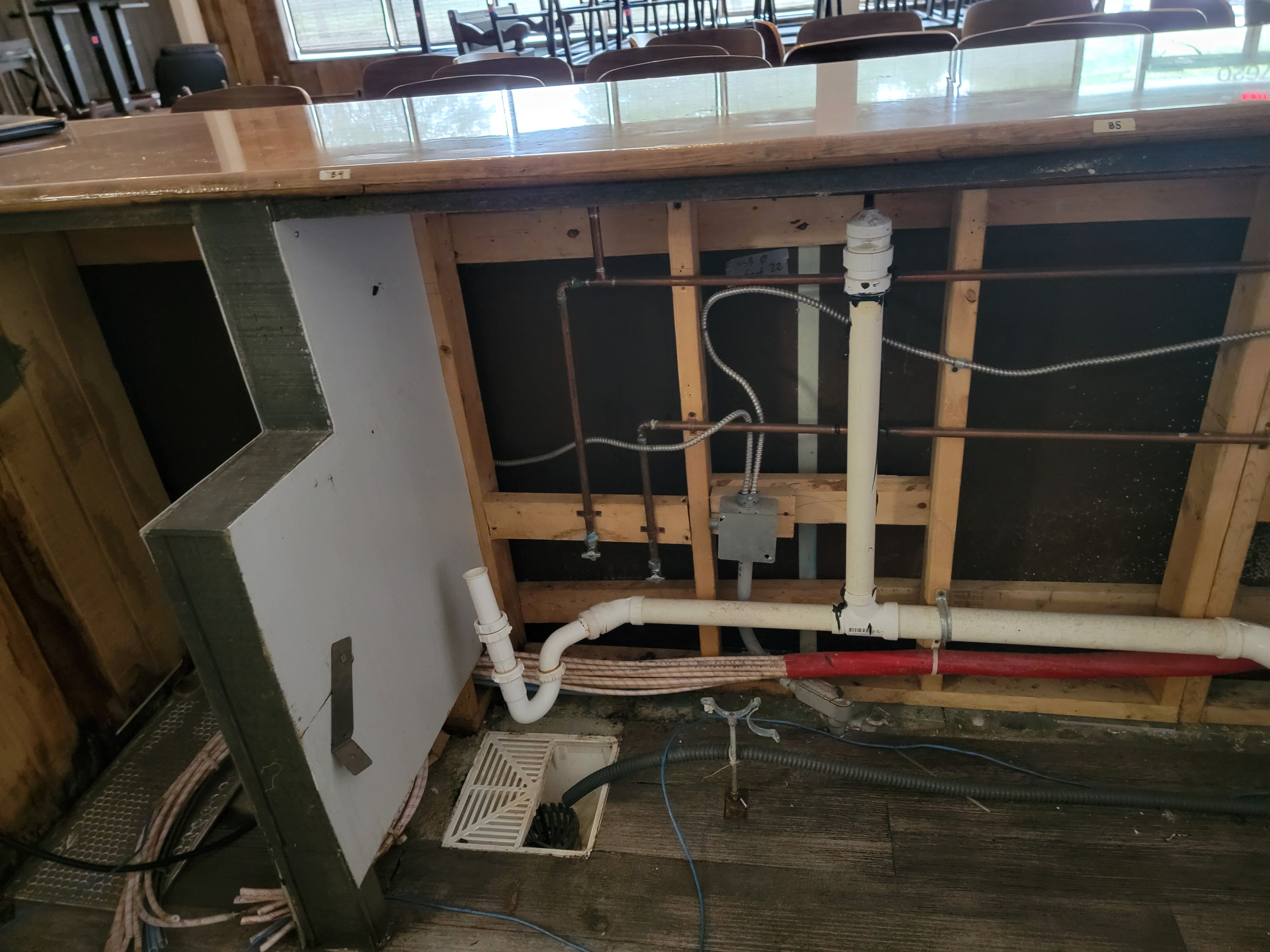 An open stud wall behind a service bar with copper water lines, white PVC waste and vent piping, flexible electrical conduit, a junction box, and a floor drain set in the deck below.