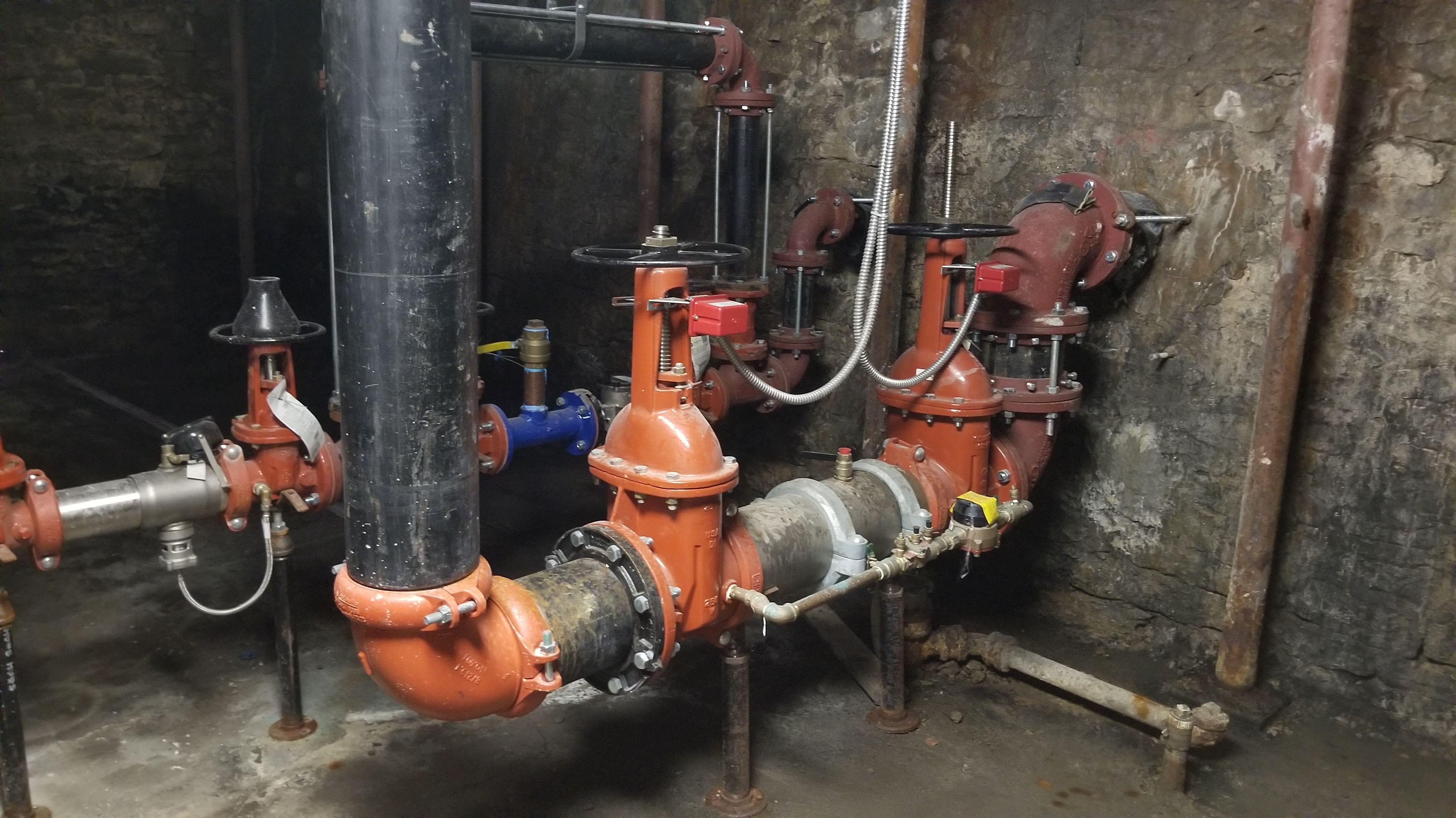 Red fire protection piping with gate valves, a backflow preventer, and tamper switches entering a stone-walled basement, with a blue domestic water line branching off beside a black standpipe.