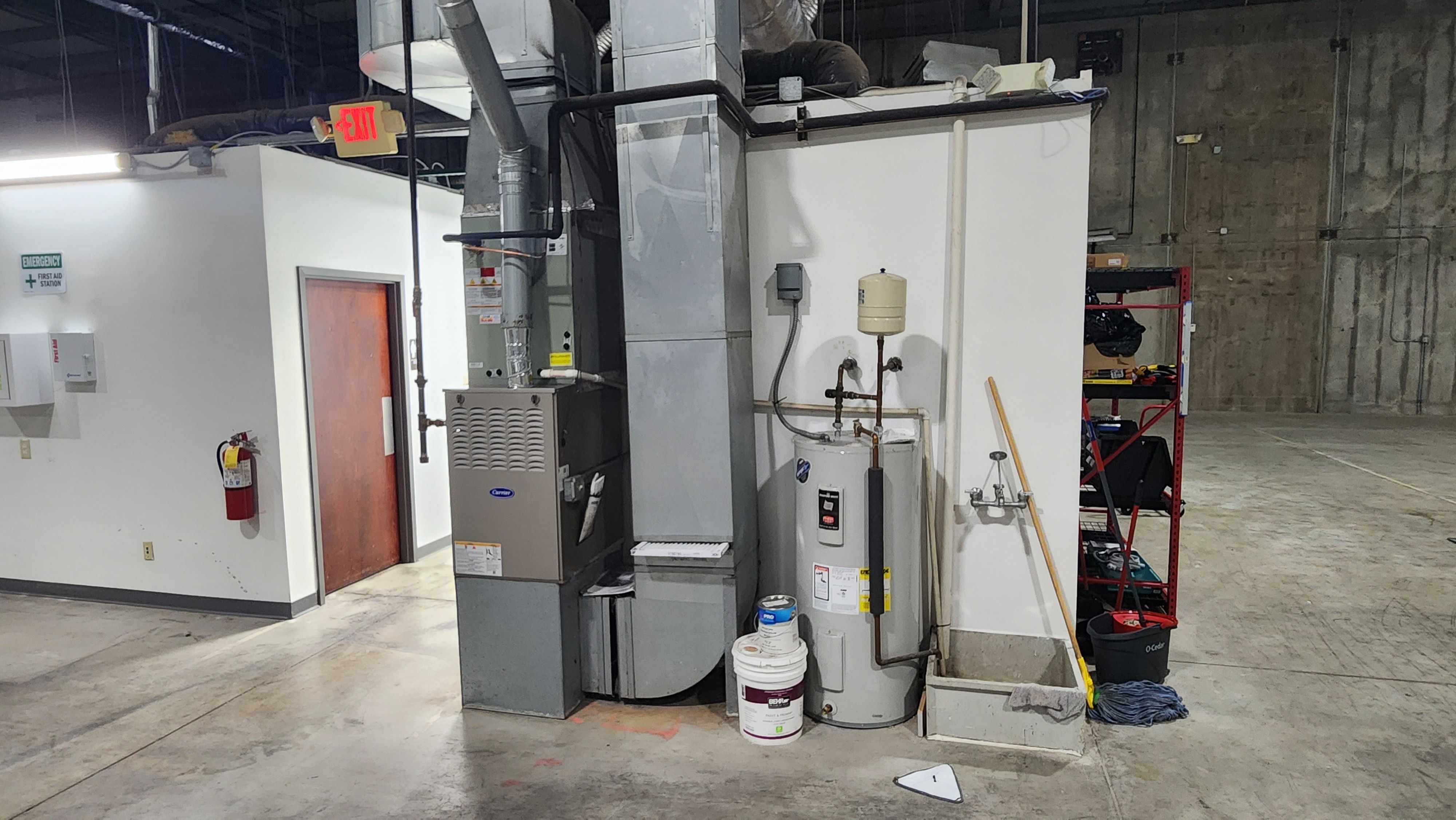 A gas furnace with a sheet metal supply plenum and return duct standing beside an electric storage water heater and its thermal expansion tank, in the corner of an open warehouse space.