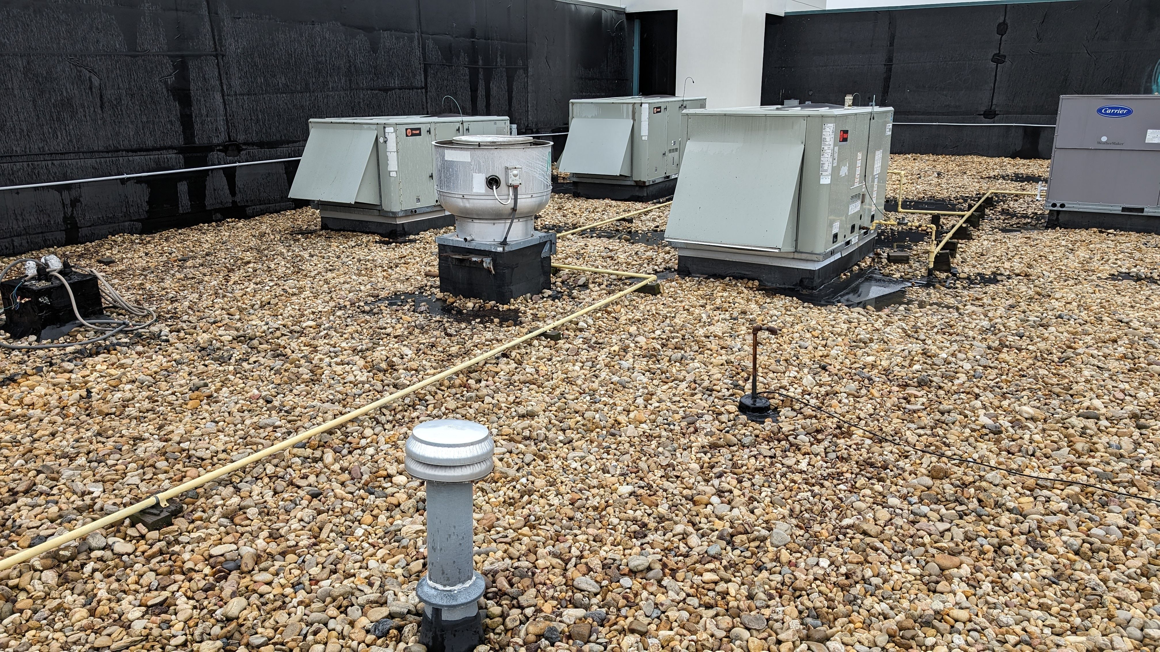 Three packaged rooftop HVAC units, a round exhaust fan, and a plumbing vent set on curbs across a gravel-ballasted roof, with a yellow gas line running between them.