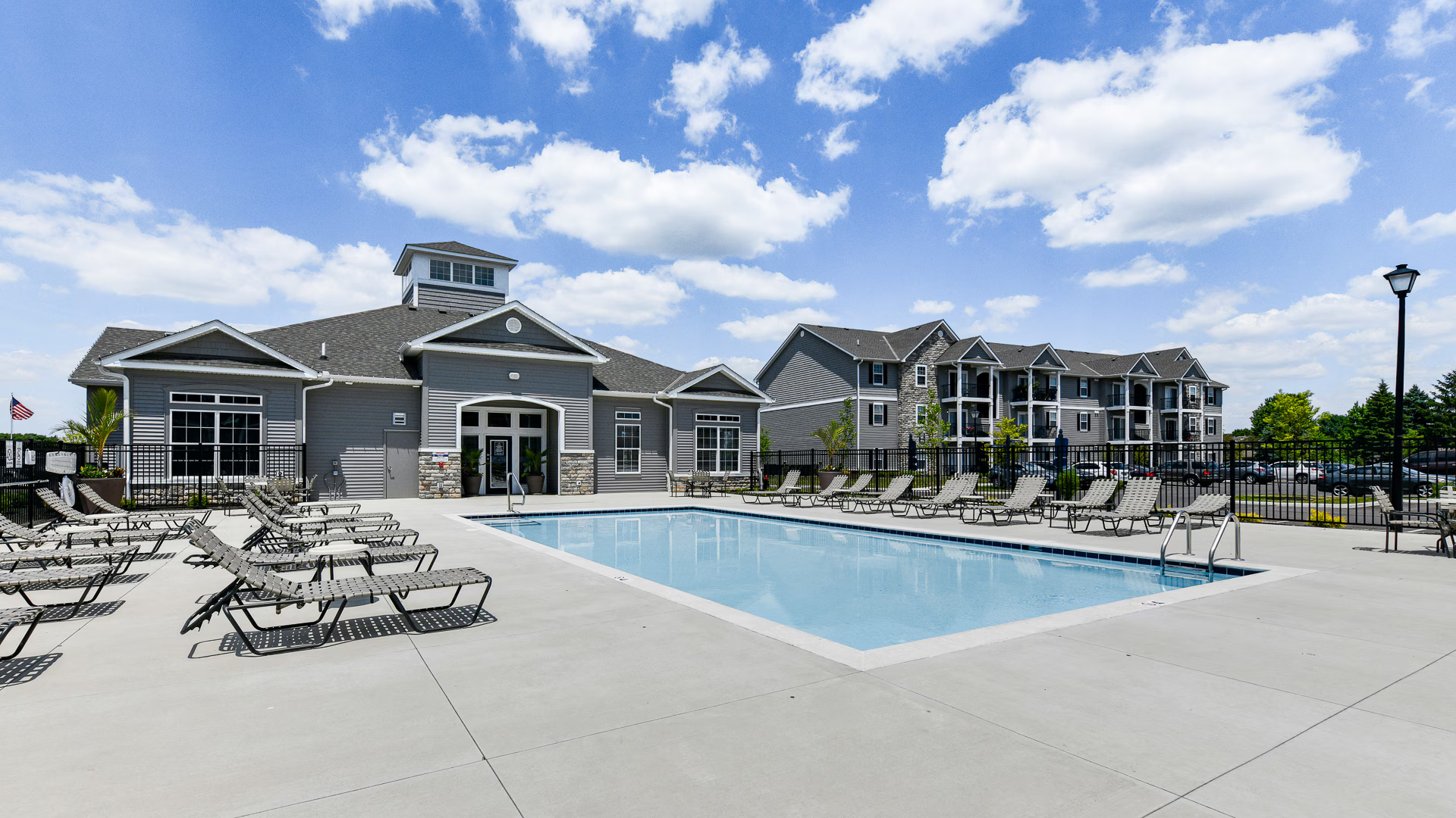 Gray apartment complex with clubhouse and outdoor swimming pool surrounded by lounge chairs on a sunny day.